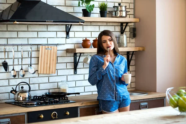 Happy beautiful brunette woman is in the kitchen with a cup of tea or coffee in blue pajamas. Early morning rise is a good habit — Stock Photo, Image