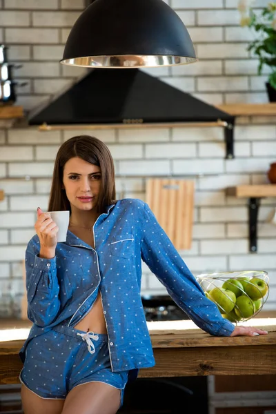 Primer plano retrato de una hermosa joven con taza de té o café en pijama azul en la cocina. Bebidas vigorizantes por la mañana para energía todo el día —  Fotos de Stock