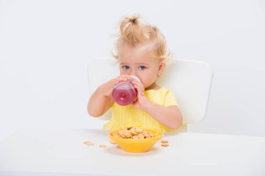 Sevimli küçük bebek kız 1 tahıl gevreği yeme ve içme suyu veya Komposto bir şişe beyaz arka plan üzerinde izole masada yaşında.