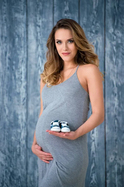 グレーの夏ドレスの美しい妊婦が木製の背景と彼女の手でベビー スニーカー ブーツは小さな新生児の靴を保持しているの前に立つ — ストック写真