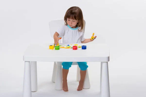 一个可爱开朗的小女孩的特写镜头画她自己的手与水粉或手指油漆在桌子上查出在白色背景 — 图库照片