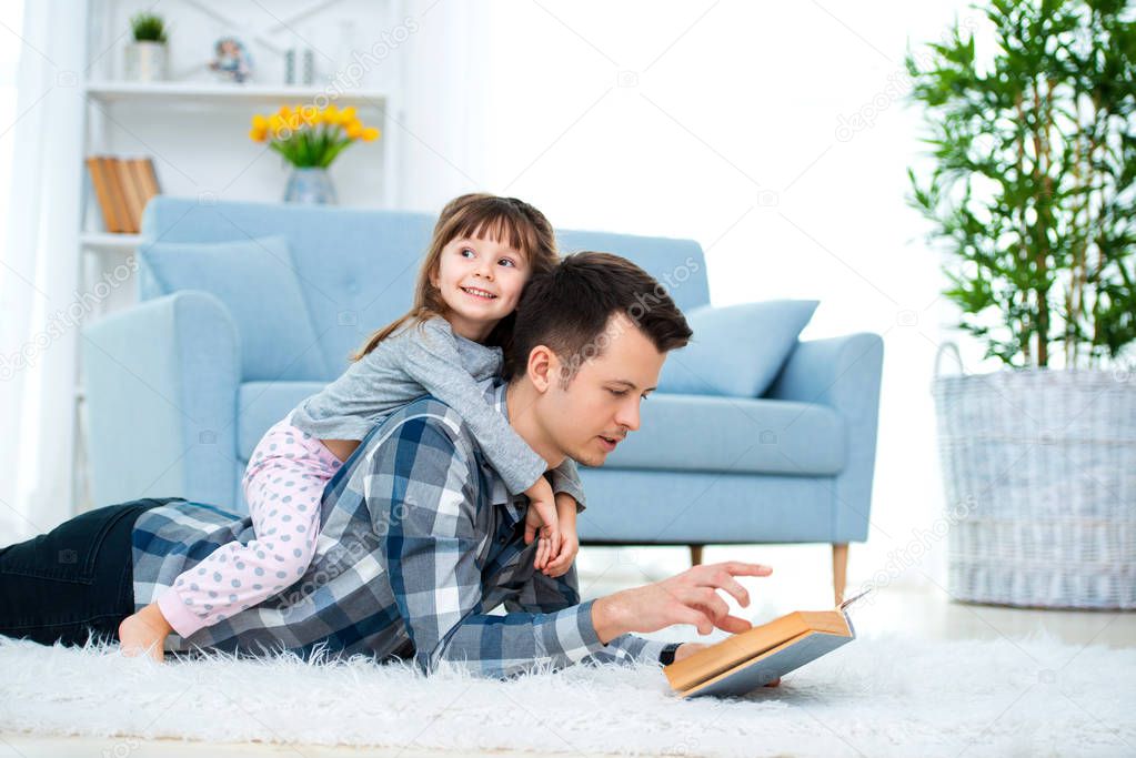 Happy family and father's day concept. Dad with daughter spending time togetherness at home. Cute little girl on dad's back lying on warm floor.