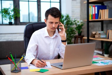 Ofiste, hücre ve alır notlar tarafından konuşan genç girişimci adam otururken dizüstü kullanarak telefonu konuşurken başarılı iş adamı.