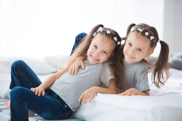 两个可爱的微笑的女孩姐妹双胞胎坐在房间的床上 — 图库照片