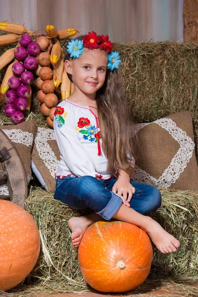 Χαμογελώντας ξανθιά κοπέλα με μακριά μαλλιά σε ένα ζωηρόχρωμο στεφάνι Ουκρανικά και το κεντημένο κάθεται σε θημωνιές. Φθινοπωρινή διακόσμηση, συγκομιδή με κολοκύθια — Φωτογραφία Αρχείου