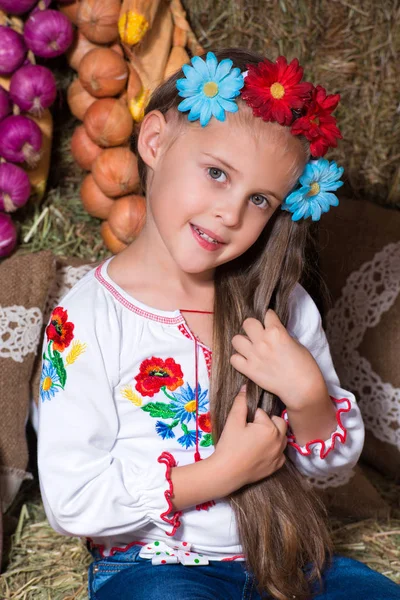 Sorrindo menina loira com cabelos longos em uma grinalda ucraniana colorida e em bordado está sentado em palheiros. Decoração de outono, colheita com abóboras — Fotografia de Stock