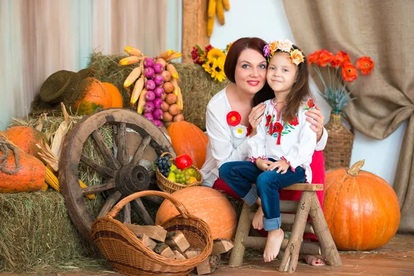 Симпатичные, улыбающиеся мать и дочь в красочном украинском венке и в вышитых сидят на стогах сена. Осенний декор, сбор урожая тыквами — стоковое фото