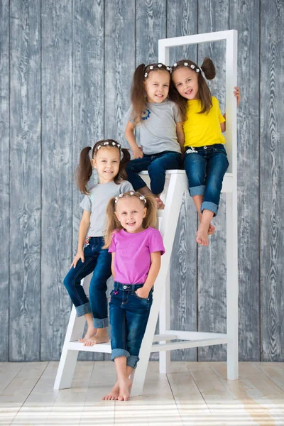 Cuatro hermosas niñas alegres hermanas gemelas triples en jeans están sentadas en una escalera delante de un fondo de pared de madera en el estudio —  Fotos de Stock