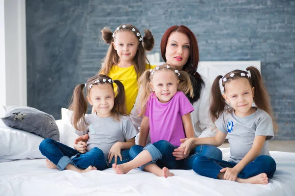 Große freundliche Familie, viele Kinder: Mutter und vier hübsche fröhliche Mädchen, dreifache Zwillingsschwestern, die auf einem Bett vor einer grauen Ziegelwand sitzen — Stockfoto