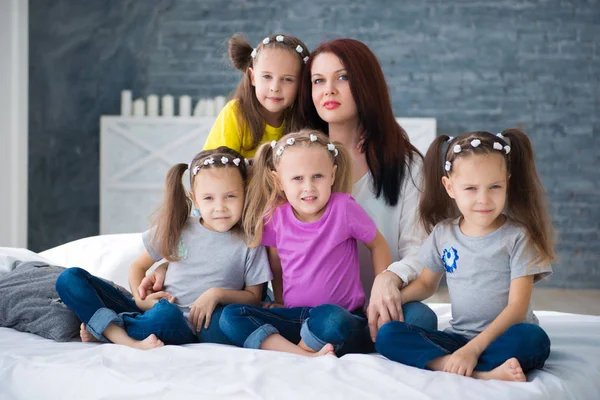 Grande família amigável, muitas crianças: mãe e quatro meninas muito alegres triplas irmãs gêmeas sentadas em uma cama contra uma parede de tijolo cinza — Fotografia de Stock