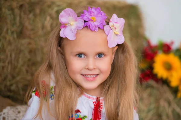 Smiling blond girl with long hair in a colorful Ukrainian wreath and in embroidered is sitting on haystacks. Autumn decor, harvest with pumpkins — Stock Photo, Image