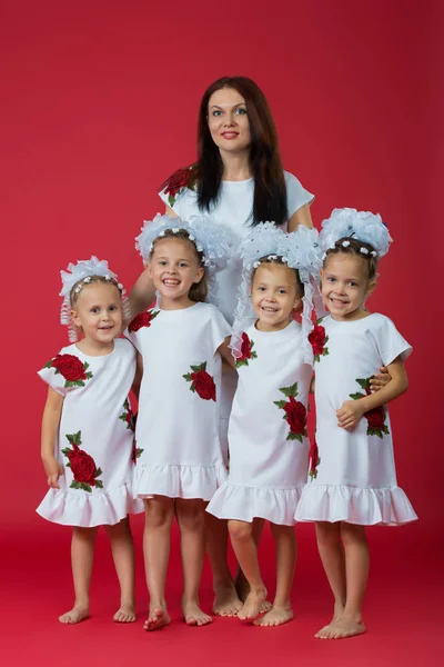 Feliz gran familia de madre y cuatro hijas niñas en vestidos blancos bordados sobre un fondo rojo liso en el estudio —  Fotos de Stock