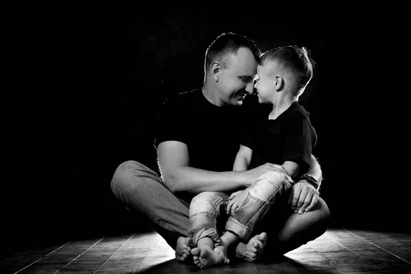 Vater nimmt Sohn in den Arm und umarmt ihn. Mann und Junge sitzen zusammen vor schwarzem Hintergrund. glückliche Vaterschaft und Familienliebe — Stockfoto