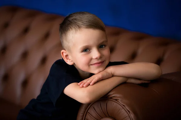 Menino bonito pré-escolar com cabelo loiro senta-se no sofá, olha para a câmera e sorri — Fotografia de Stock