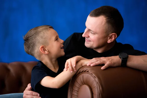 Father and little son are sitting on a leather sofa, smiling and talking to each other. Happy fatherhood and family love. Family portrait — Stock Photo, Image