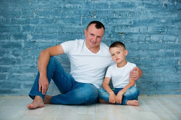 Pai e filho em jeans casuais e camisetas brancas estão sentados em um piso de madeira quente perto de um fundo de parede de tijolo cinza — Fotografia de Stock