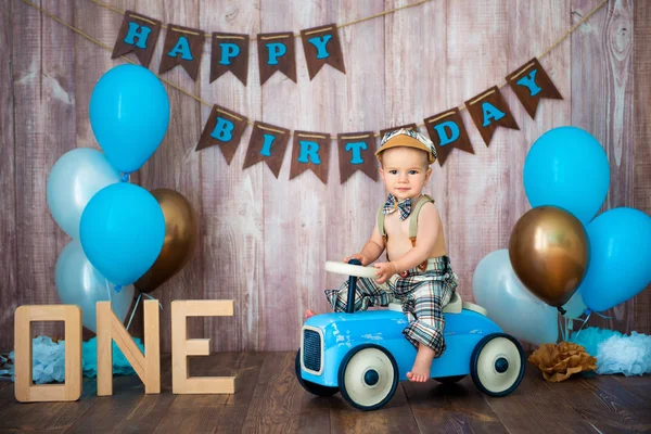 Ein kleiner Junge im Retro-Kostüm mit Hosenträgern und Mütze sitzt auf einem hölzernen Auto. Kinderfest mit Luftballons zum Geburtstag, 1 Jahr — Stockfoto