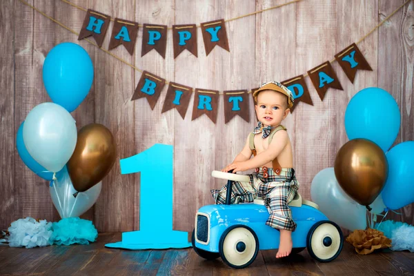 Ein kleiner Junge im Retro-Kostüm mit Hosenträgern und Mütze sitzt auf einem hölzernen Auto. Kinderfest mit Luftballons zum Geburtstag, 1 Jahr — Stockfoto