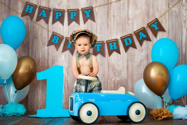 Ein kleiner Junge im Retro-Kostüm mit Hosenträgern und Mütze sitzt auf einem hölzernen Auto. Kinderfest mit Luftballons zum Geburtstag, 1 Jahr — Stockfoto