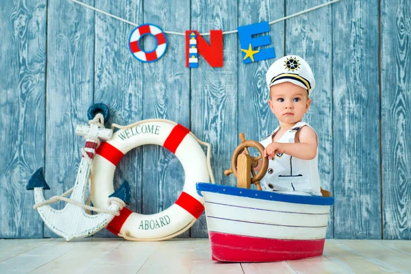 Фотосессия для мальчика года. Маленький морской капитан, моряк на игрушечном корабле с рулем. Морской якорь и спасательный круг на сером деревянном фоне — стоковое фото