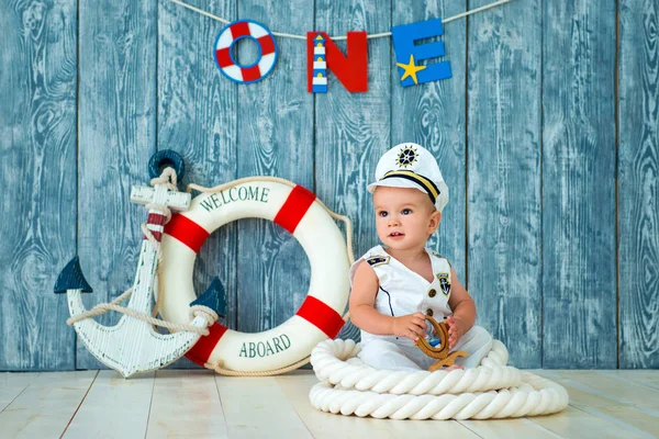 Servizio fotografico per un ragazzo di un anno. Piccolo capitano di mare, marinaio su una nave giocattolo con volante. Ancora di mare e salvagente su sfondo grigio in legno — Foto Stock