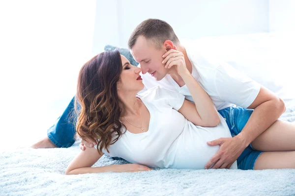 Cute pregnant couple woman in jeans overalls and a man lying on the bed near the window in a bright room — Stock Photo, Image