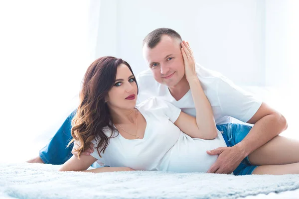 Cute pregnant couple woman in jeans overalls and a man lying on the bed near the window in a bright room