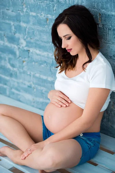 Donna incinta indossa tuta jeans e una t-shirt bianca su sfondo grigio muro di mattoni in studio interno — Foto Stock