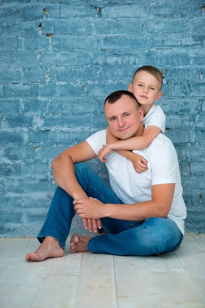 Vater und Sohn in lässigen Jeans und weißen T-Shirts sitzen auf einem warmen Boden und umarmen sich an einer grauen Ziegelwand — Stockfoto