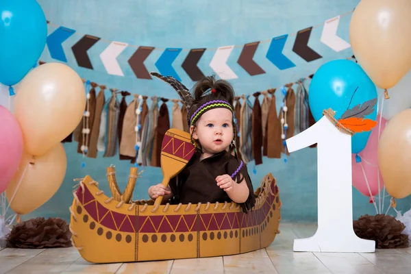 Little cute girl in costume of Apache Indian chief and in a feather headdress sits in canoe. Childrens holiday First birthday and smash cake 1 year
