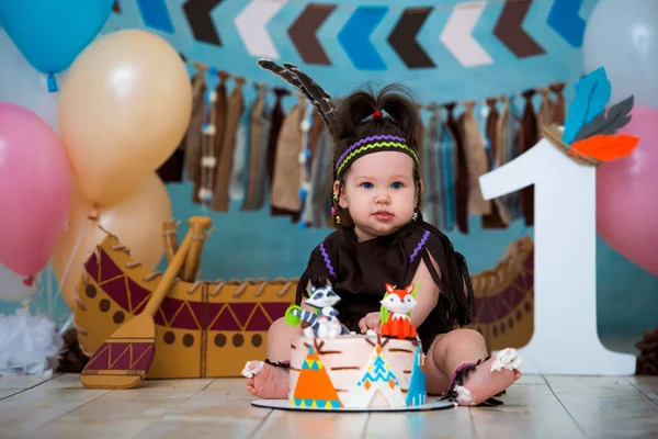 アパッチ インディアン チーフのコスチュームと羽のヘッドドレスでかわいい女の子は座っているし、味が甘いケーキ。最初誕生日の 1 年とスマッシュのケーキ — ストック写真