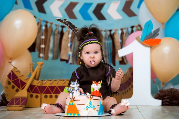 Little cute girl in costume of Apache Indian chief and in a feather headdress sits and tastes a sweet cake. First birthday 1 year and smash cake
