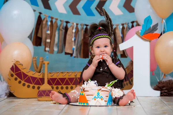 Мила дівчинка в костюмі Apache індіанського вождя і в це головний убір сидить і смак солодкий брикету. Перша 1 рік і розбити торт до дня народження — стокове фото