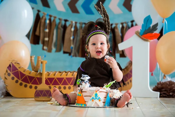 Little cute girl in costume of Apache Indian chief and in a feather headdress sits and tastes a sweet cake. First birthday 1 year and smash cake