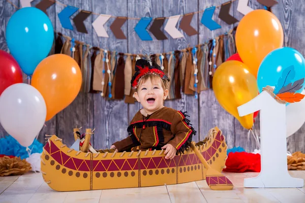 Маленький милый мальчик в костюме индейского вождя апачей сидит в каноэ. Детский праздник, с днем рождения 1 год, украшение фотозоны — стоковое фото