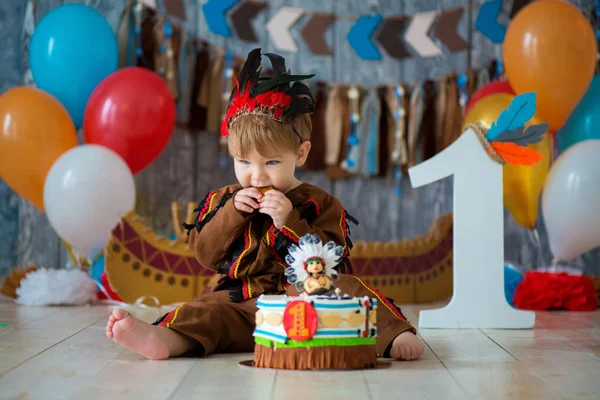 little cute boy in costume of American Indian Apache Chief sits and smash cake. Children\'s holiday, happy Birthday 1 year, photo zone decoration