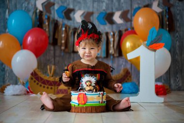 little cute boy in costume of American Indian Apache Chief sits and smash cake. Children's holiday, happy Birthday 1 year, photo zone decoration clipart
