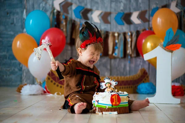 Photo shoot smash the cake for 1 year in the style of Indian chief. Little emotional boy with a tomahawk in costume of Apache and in feather headdress