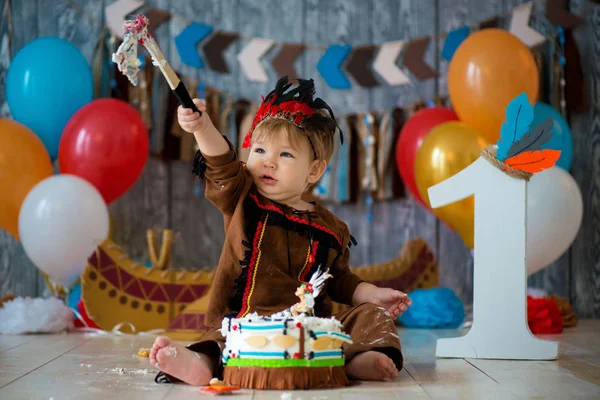 Photo shoot smash the cake for 1 year in the style of Indian chief. Little emotional boy with a tomahawk in costume of Apache and in feather headdress