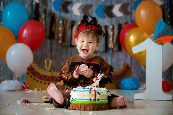 Kleiner süßer Junge im Kostüm eines amerikanischen indianischen Apachen-Häuptlings sitzt und zerschlägt Kuchen. Kinderurlaub, Happy Birthday 1 Jahr, Fotozonendekoration — Stockfoto