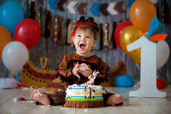 Kleiner süßer Junge im Kostüm eines amerikanischen indianischen Apachen-Häuptlings sitzt und zerschlägt Kuchen. Kinderurlaub, Happy Birthday 1 Jahr, Fotozonendekoration — Stockfoto