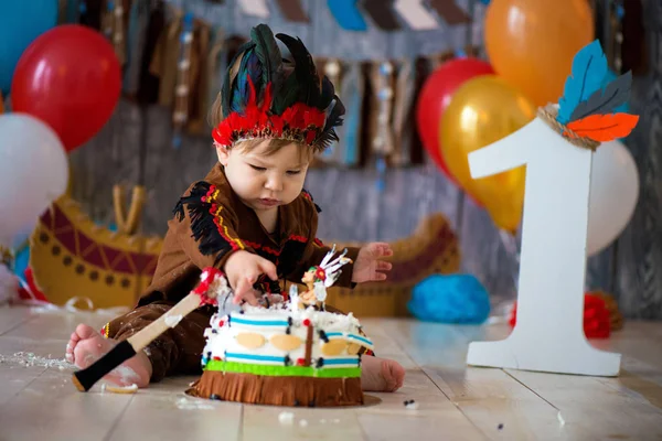 Photo shoot smash the cake for 1 year in the style of Indian chief. Little emotional boy with a tomahawk in costume of Apache and in feather headdress