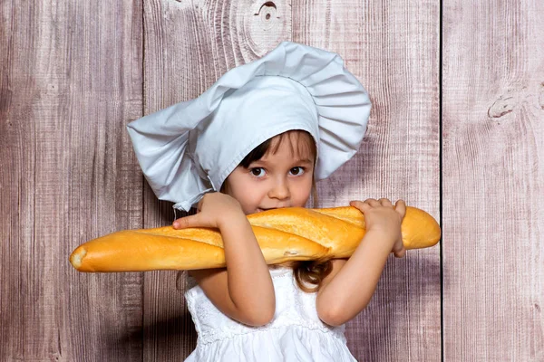 Nahaufnahme eines kleinen lächelnden Mädchens mit Kochmütze und frischem Baguette in den Händen — Stockfoto