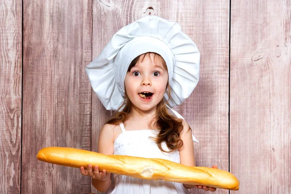 Close-up portrait of a little smiling girl in a cooking cap with a fresh baguette in her hands