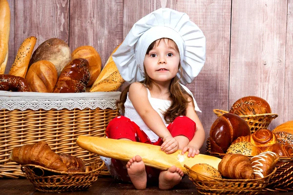 Schattig klein lachende meisje in een chef GLB zitten met een stokbrood in haar handen in de buurt van de rieten mand met broodjes en bakkerijproducten — Stockfoto