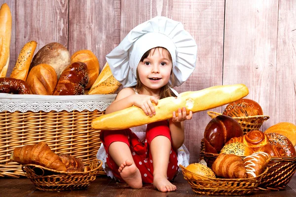 Menina sorridente bonito em um boné de chef sentado com uma baguete em suas mãos perto da cesta de vime com pães e produtos de padaria — Fotografia de Stock