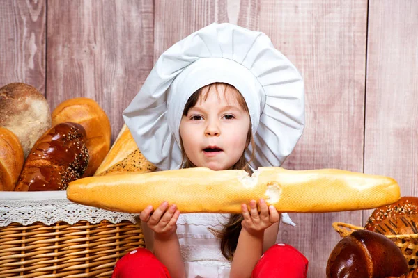 Nettes kleines lächelndes Mädchen in Kochmütze mit Baguette in der Hand neben dem Weidenkorb mit Brötchen und Backwaren — Stockfoto