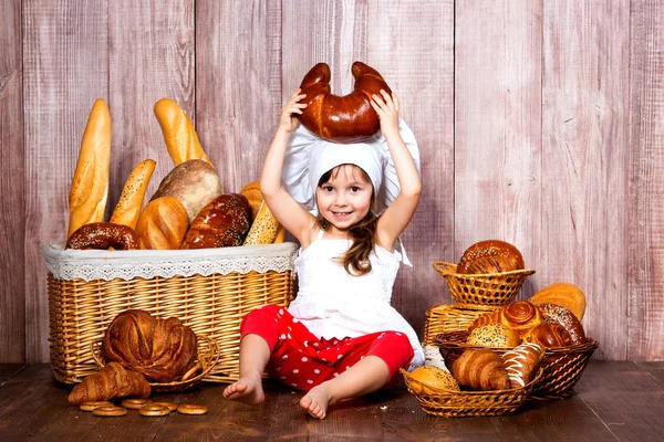 Positiv lächelndes kleines Mädchen mit Kochmütze sitzt mit einem Bagel in der Hand neben dem Weidenkorb mit Brötchen und Backwaren — Stockfoto