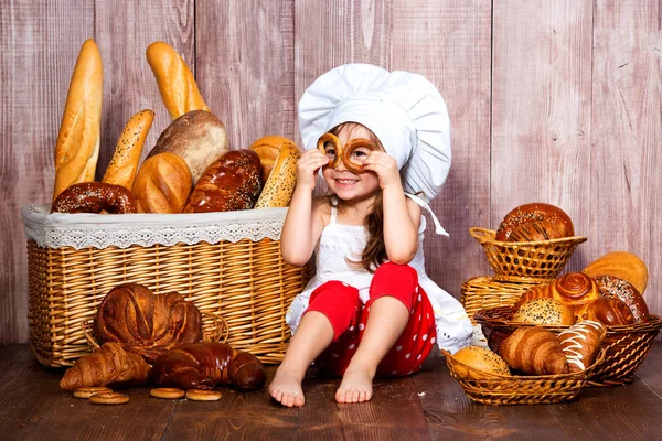一个戴着厨师帽的积极的微笑的小女孩在柳条篮子附近做百吉饼眼镜, 里面有面包卷和面包店的产品 — 图库照片