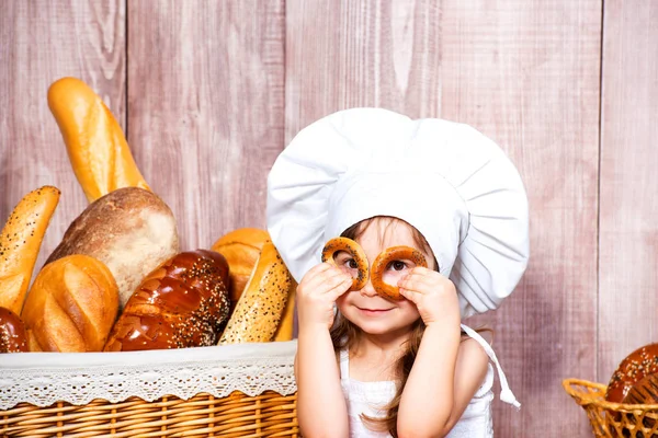 Positieve beetje lachende meisje in een cook cap maakt bagels bril in de buurt van een rieten mand met broodjes en bakkerijproducten — Stockfoto